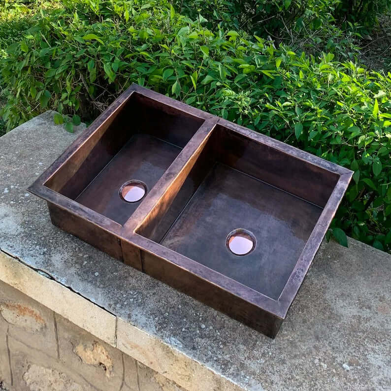 Handmade copper double vessel sink for kitchen, showcasing elegant undermount design against natural greenery.