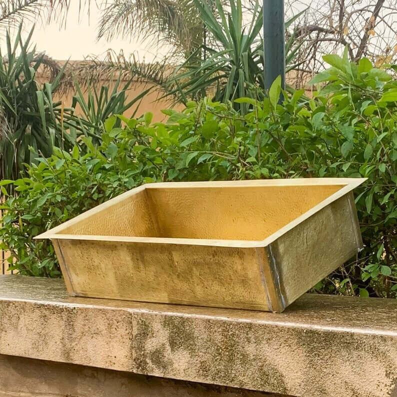 Handmade golden brass sink placed on a stone countertop with greenery in the background.