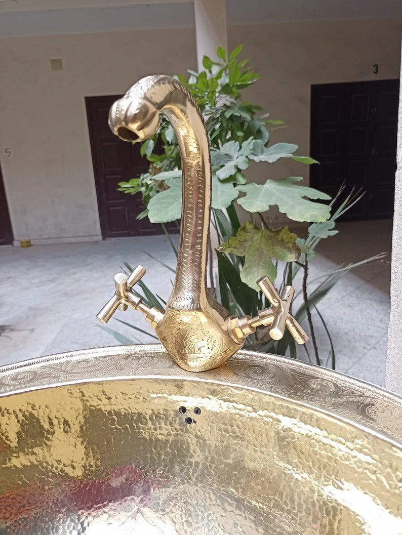 Stylish copper basin faucet with ornate design and two handles, surrounded by greenery in a modern setting.