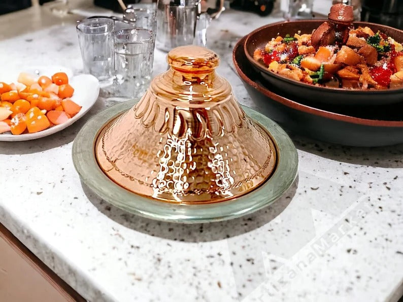 Handcrafted Moroccan copper tagine with lid on serving tray, alongside a bowl of colorful fruit on a kitchen counter.