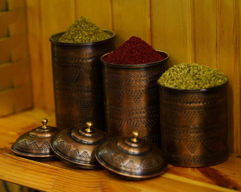Handmade copper spice jars in a rustic kitchen, filled with various spices and adorned with intricate designs.