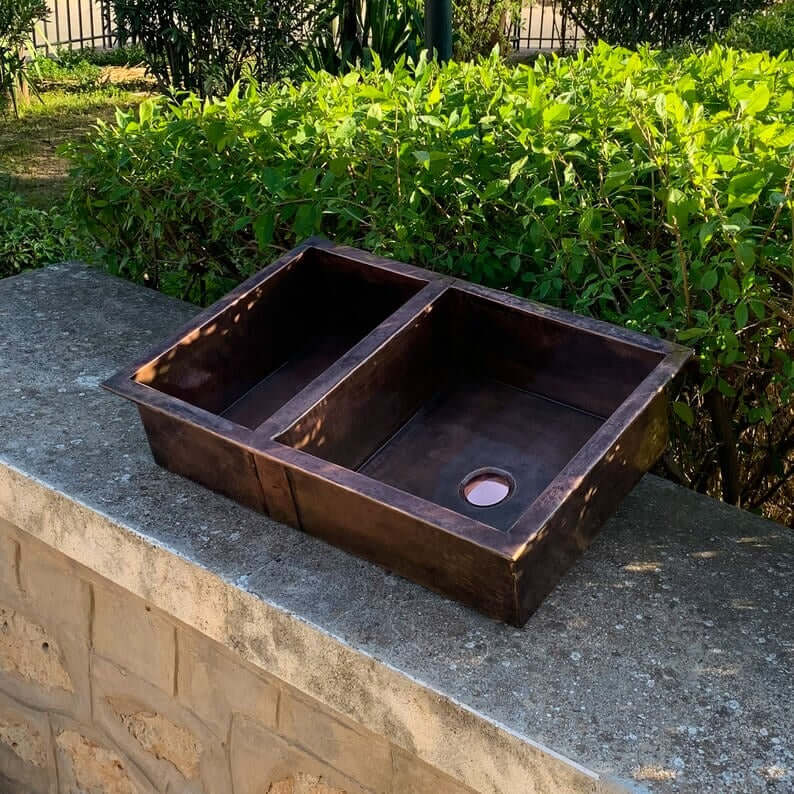 Cooper handmade double vessel sink for kitchen, crafted from high-quality copper, displayed outdoors on a stone ledge.