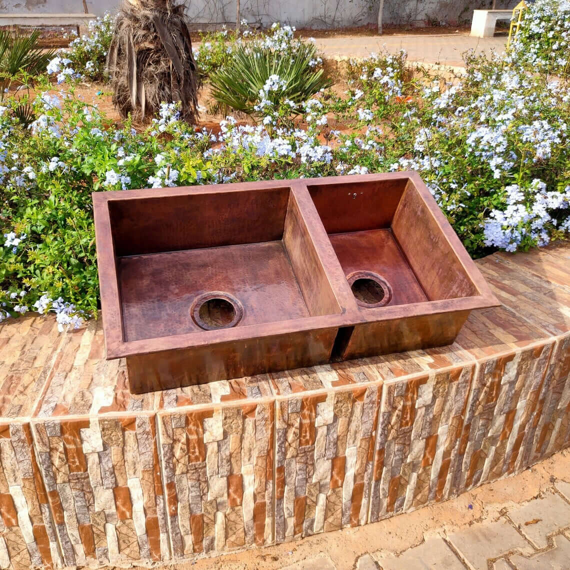 Handmade double copper sink on a stone base surrounded by flowers, perfect for farmhouse kitchens.