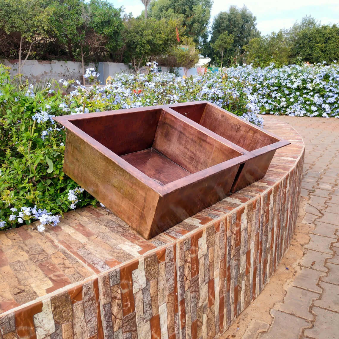 Handmade double copper sink displayed outdoors among flowers, showcasing its rustic design and craftsmanship.