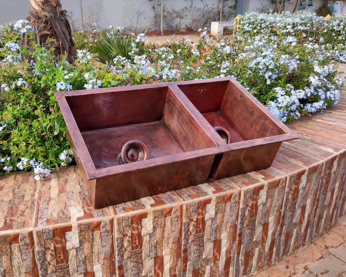 Handmade double copper sink displayed outdoors, highlighting its rustic elegance in a garden setting.