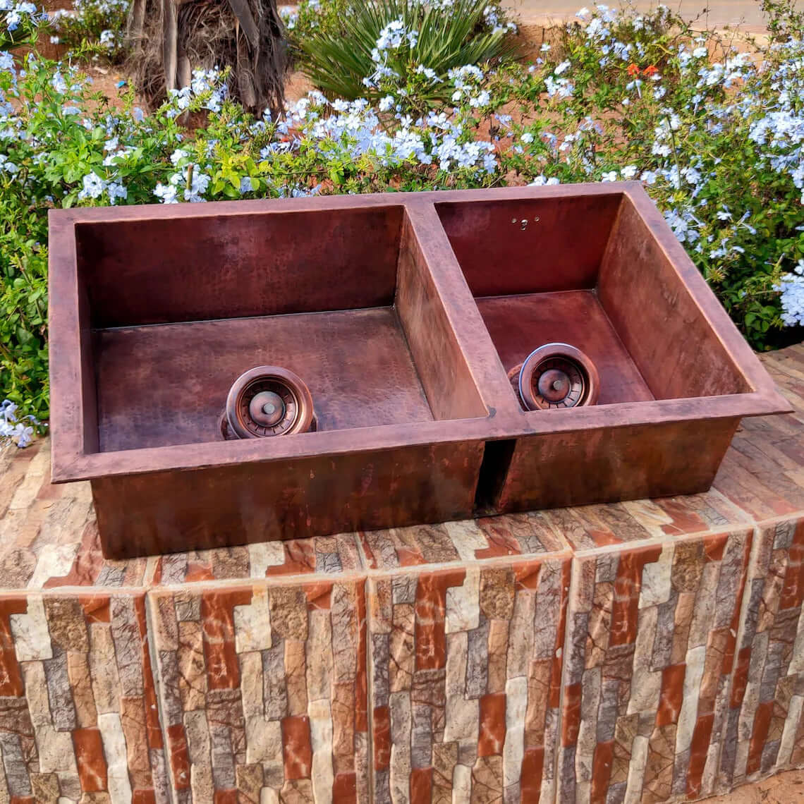 Handmade double copper sink with hammered finish, designed for a farmhouse kitchen, surrounded by flowers.