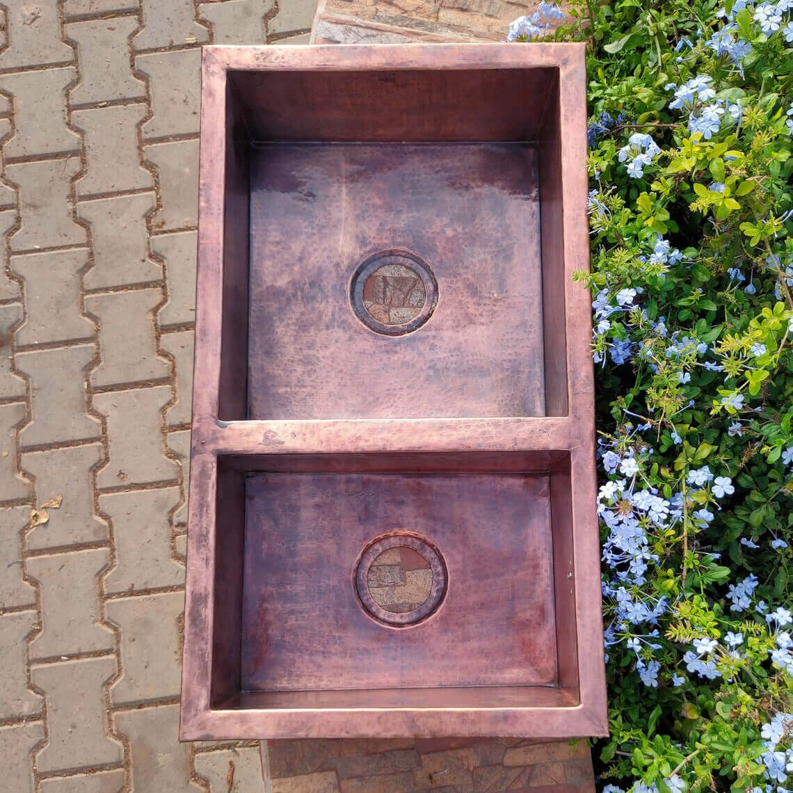 Handmade hammered double copper sink, showcasing Moroccan craftsmanship, surrounded by flowers and outdoor stone flooring.