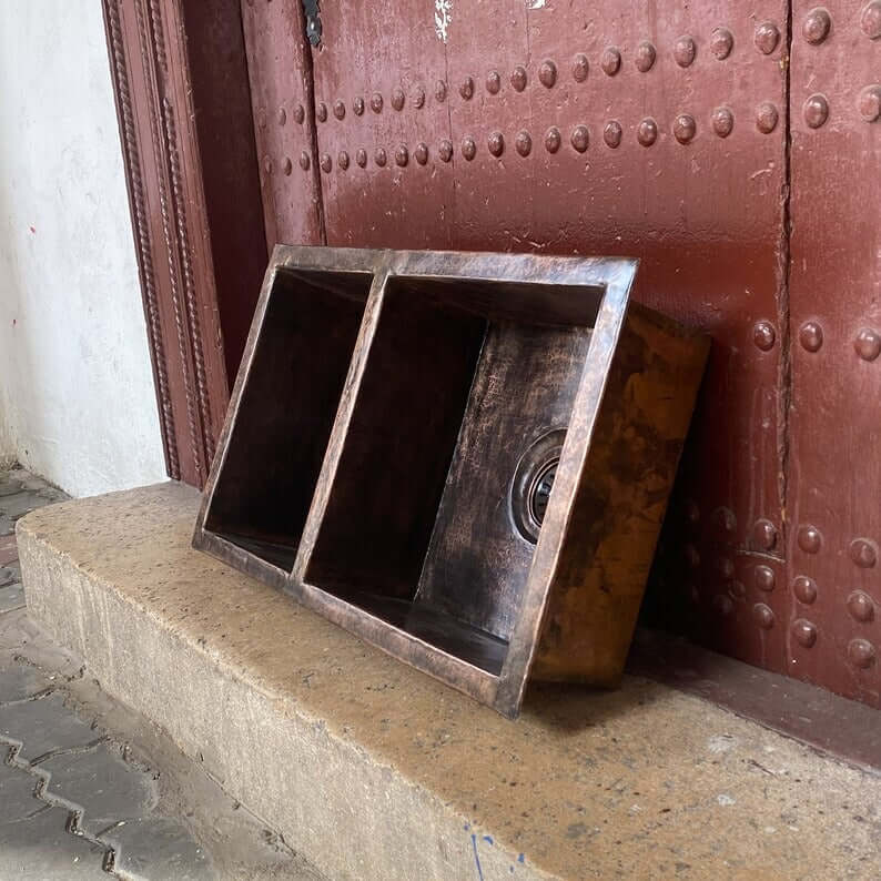 Handmade double vessel copper sink displayed against a textured wall, showcasing its unique design and craftsmanship.
