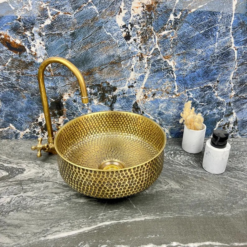 Hand-hammered brass round vessel sink with drain, set against a marble background, showcasing luxury and craftsmanship.