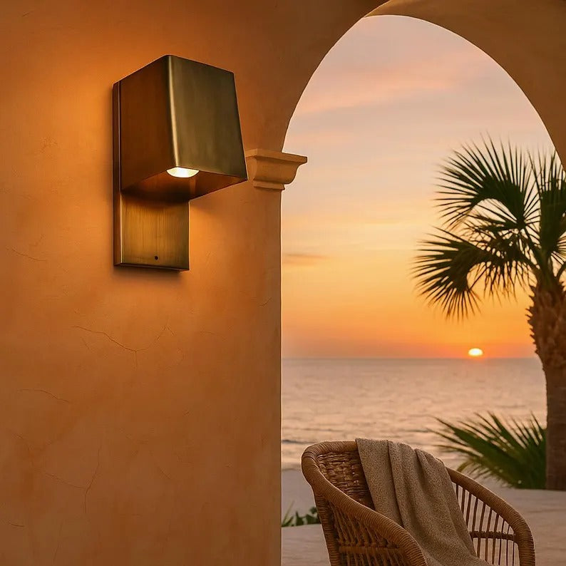 Handcrafted copper wall sconce casting warm light at sunset by the ocean, blending modern design with nature.