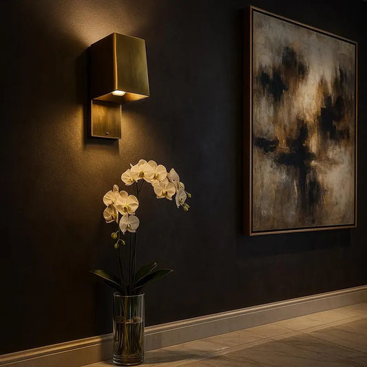 Modern brass wall sconce illuminating dark wall with white orchids in vase and abstract art.