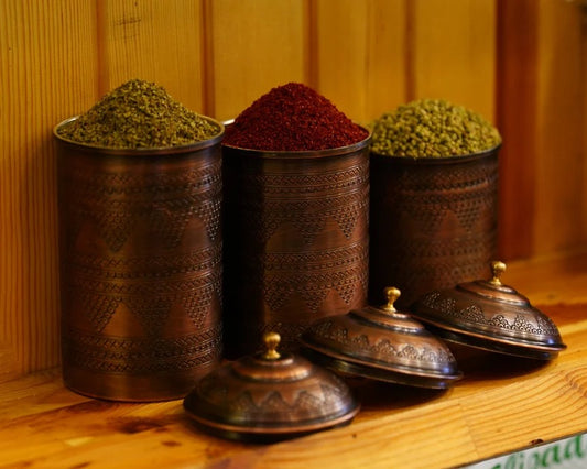 Handmade copper spice containers with colorful spices, featuring intricate designs and elegant lids, displayed on a wooden shelf.
