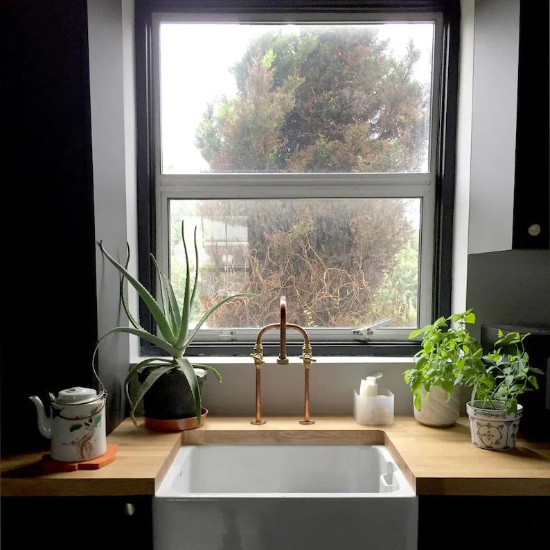 Handmade copper and brass deck mount faucet above a farmhouse sink with potted plants in a cozy kitchen setting.