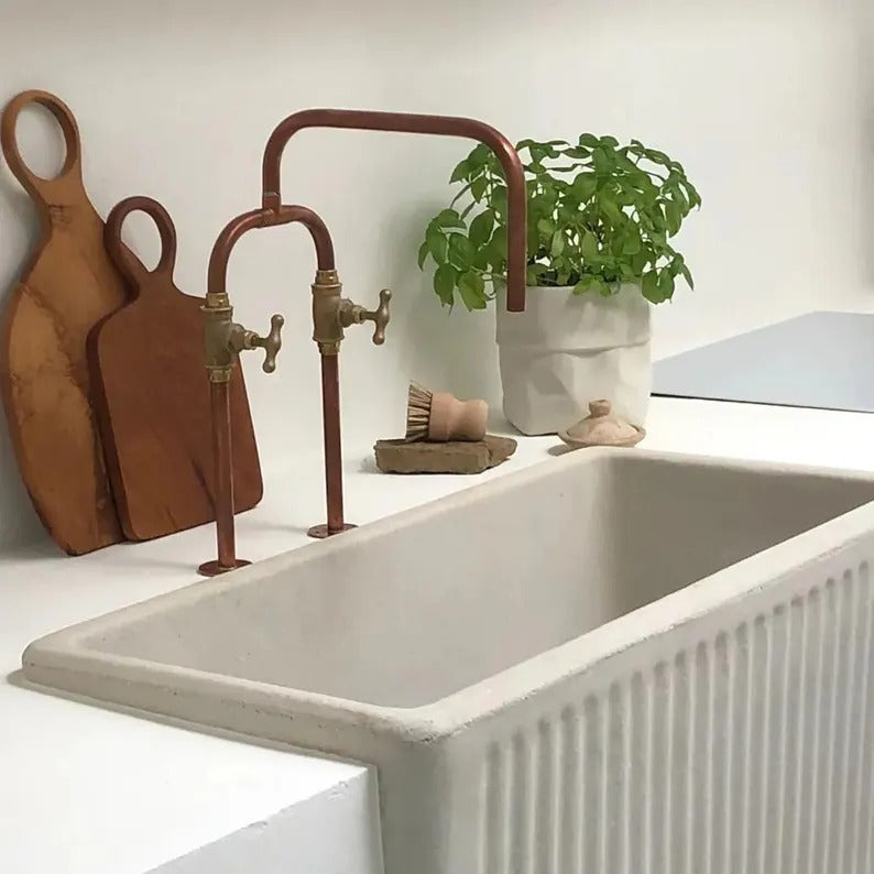 Handmade copper and brass faucet above a white farmhouse sink with green plant and wooden cutting boards.