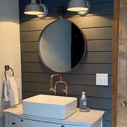Handmade copper faucet above white vessel sink in stylish bathroom with dark wood paneling and round mirror.